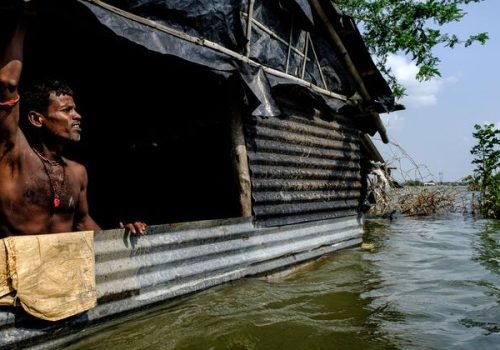 Ocean Image Bank/Dipayan Bose
                          Um homem está na porta de sua casa inundada, de telhado de zinco, em uma vila costeira na Índia